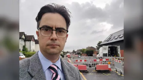 LDRS A man with dark hair in a blue shirt, grey blazer, blue and pink tie and glasses. He is standing in front of road works.