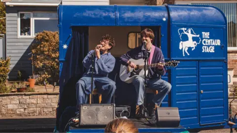 Ciaran Gallen Two men sat on a horsebox-turned-stage, one singing and one playing the guitar