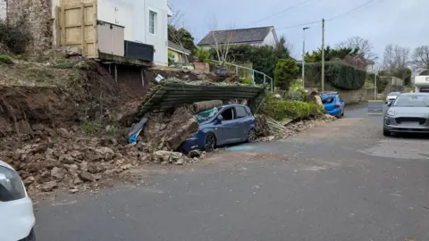 A wall has collapsed onto a parked car on a road in Bideford. The car is blue. Another car - which is a lighter shade of blue - parked further down the road has also been damaged. Houses line the road.