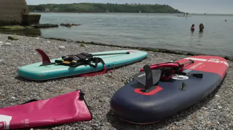 Two paddleboards are resting on a pebbly beach. The sea in the background is calm. Two women are stood in the water. 
