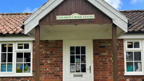 A close up of a white door that is the entrance to a shop. Above the door is an awning with a yellow and green sign that reads "Norfolk Children's Book Centre". The building is red brick and one storey with two windows either side of the door. 