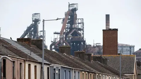 Getty Images The first floor and roofs of a row of a street of colourful terraced homes can be seen across the lower half of the picture. The skyline behind them is dominated by a grid of pipes and metalwork which make up the two blast furnaces at Tata steel. 