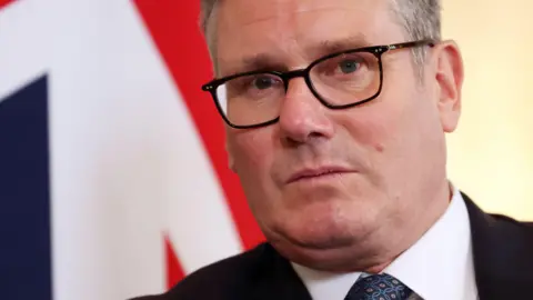 EPA Sir Keir Starmer wearing a shirt, tie and suit pictured with a union jack behind him.