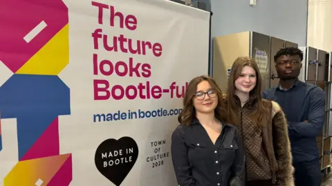 Three students - two girls one boy - stand in front of a sign at Hugh Baird college, Bootle which says" The Future is Bootle - Full"