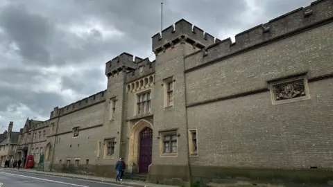 A view of the exterior of County Hall in Ipswich. The building is made of large grey bricks and has a similar look to a castle. People can be seen walking past. 