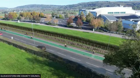 An artist's mockup of the new cycleway with the cycle path painted green. There is a footpath on one side and a two-lane road, bordered by hedges and a grassy area, on the other. A white building and car park can be seen in the background.