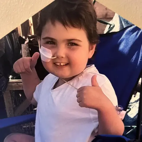 Mark Bissett Charly - A child sits in a blue folding chair wearing a white shirt, holding both hands up with thumbs extended in a thumbs‑up gesture. Outdoor wooden structures and various items are visible in the background.