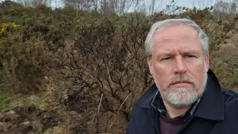 Durham County Councillor Mark Wilkes. He has short white hair and a trimmed beard. He has blue eyes and is wearing a blue coat, over a purple jumper. He is looking at the camera with a serious expression. Behind him are some gorse bushes.