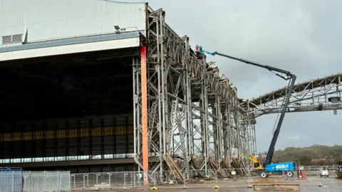 The hangars looking bare and skeletal ready for construction to begin. A person is operating a blue cherry picker in the distance.