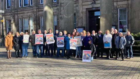 A view of a group of campaigners outside Macclesfield Town Hall. A number of the group are holding signs, including one that says "save our trees". Most of the campaigners are wearing large coats as it was a cold day.