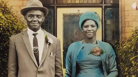 A man and a woman standing outside a house in a vintage photograph, with the woman wearing a blue dress and carrying a handbag.