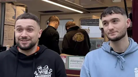 The brothers - both with short dark hair and short beards - stand in front of the serving hatch of the tram carriage where they sell the potatoes. Both are wearing hoodies and two people are in the background preparing the spuds.