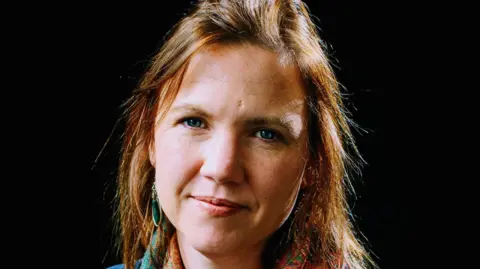 Jade Levell A professional headshot of Dr Jade Levell. She has ginger hair and blue eyes and is sat in front of a black background, smiling at the camera.