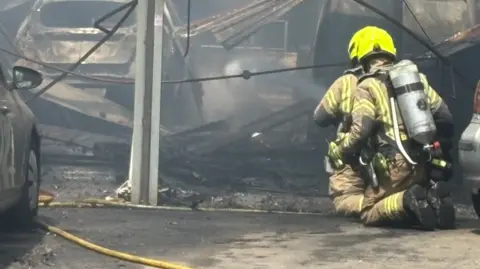 Hertfordshire Fire and Rescue Service A firefighter wearing bright yellow protective headgear and full firefighting equipment is kneeling on the ground facing the damaged building. The firefighter is wearing a breathing apparatus cylinder on their back and appears to be assessing the scene or tackling remaining hotspots. Another firefighter is in front of them.