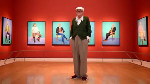 PA Media David Hockney stands in brown trousers, a green cardigan in a gallery. His 'still-life' portraits are hung on the orange walls behind him.