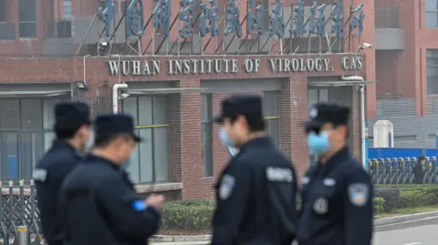 Four security guards wearing face masks, whose figures are slightly blurred because of the camera's focus, stand in the foreground of the picture. Behind them is the Wuhan Institute of Virology with signs in English and Mandarin