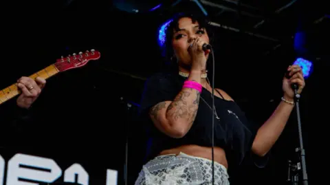 Afreine Afreine with long dark hair, wearing a black top and grey skirt. She has a pink wristband on her right arm, which also has tattoos. She is holding a microphone in her right hand and a microphone stand in her left. The fingerboard and neck of a brown guitar can be seen to the left.