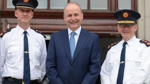 PA Media Three men smile into the camera. The man in the centre is wearing a navy suit, white shirt and light blue tie. The two other men are wearing white shirts and dark ties. They each have shoulder badges with red and yellow designs on a black background. They are also wearing dark hats with gold trim and the Garda Síochána badge.