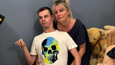 Hazel Howe Thomas Howe, with short dark hair, wearing a white T-shirt with a blue and yellow skull design, and red trousers. He is sitting on the floor with his mother behind him. She has long blonde hair and is wearing a black top. There is a large teddy bear behind them along with some cushions. There is a grey wall in the background.