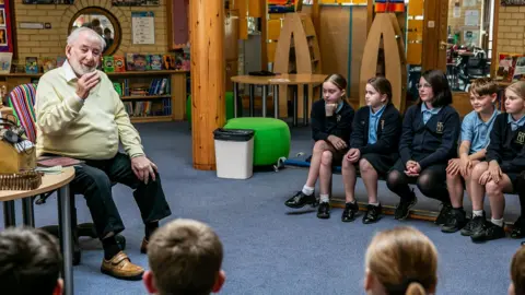 François Le Maistre sits on a chair as he talks to a group of school children sat in a semi-circle around him. Some World War Two artefacts are on the table alongside him. 