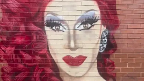 Divina De Campo A colourful mural of drag queen Divina De Campo, with red curly hair and red lipstick, with sections where the mural has been defaced by graffiti blurred out