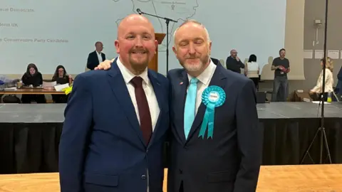 Two men smile for the camera in suits. one has a pale blue rosette and the other has a claret tie.