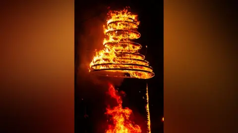 Adam Parkin Multiple rings of fire are stacked on top of each other and held aloft. The flames are bright yellow and orange in contrast to the night-time scene behind it. 