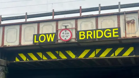BBC The close up of a railway bridge. A fluorescent yellow sign reads "Low Bridge" in capital letters. There is a circular sign between the two words that informs drivers of the bridge's height - it reads 4.2m and 13ft 9in. 