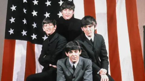 Getty Images The Beatles, in black and grey suits, shirts and polo neck jumpers, pose for a portrait in front of an American Flag