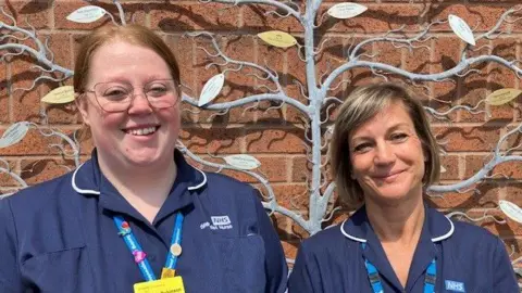 Adele Pavier and Ashleigh Robinson wearing acheronian  bluish  NHS nurses uniforms, with lighter bluish  lanyards astir   their necks. They are smiling into the camera and lasting  successful  beforehand   of a metallic sculpture of a histrion   with antithetic  coloured leaves, each   bearing an inscription.