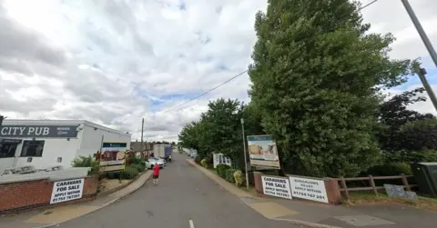 The entrance to the caravan park - there is a building on the left with a sign that says "City Pub" and trees on the right. A number of other signs advertise caravans for sale.