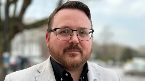 Stuart Woodward/BBC A man with short, brown hair and glasses looking serious wearing a black shirt and a white jacket but no tie.