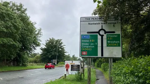 A main road, with a large road sign over the pavement to the right, showing routes to Nantwich, the M6, Stone, Stoke-on-Trent, Congleton, Chester and Wrexham