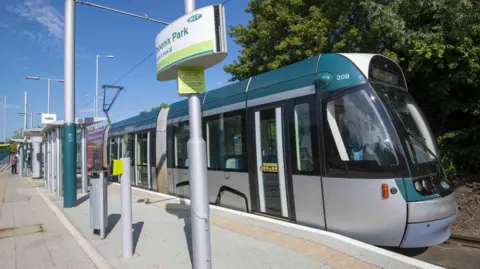 A Nottingham tram, in silvery and green livery, waits at a stop