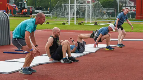Gergana Marinova Four men exercising on mats on an outdoor sports complex. They have been pictured at various points of an exercise movement which involves rolling from their back into a squat position before standing up.
