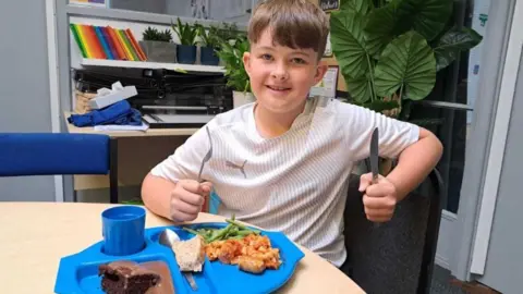 LDRS A pupil sits with a knife and fork looking like he is about to tuck into a meal and drink on a blue tray
