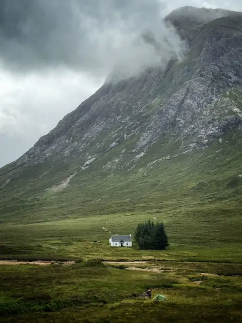 Peter Xu A small white cottage sits alone on a wide green moor beneath a towering rocky mountain. Low cloud and mist cling to the upper slopes, while a winding path leads across the landscape toward the house. In the foreground, a small tent and a person stand near a stream.