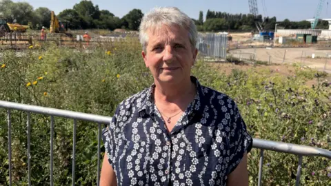 A woman wearing a black blouse with white flowers stands in front of a metal railing. Grass and a large construction site can be seen behind her.