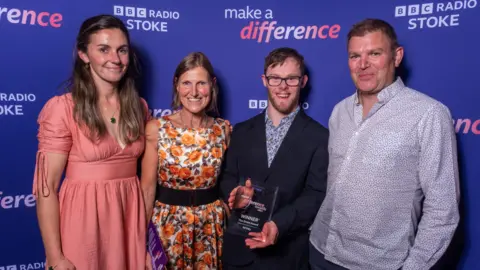 Two women and two men post for a photo, with one of the men in a black jacket holding an award