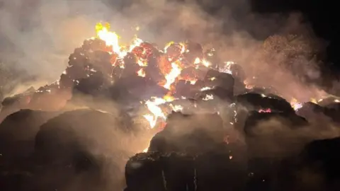 Dark large mound of hay with gold, white and orange fire at its centre and smoke plumes going up into the night sky