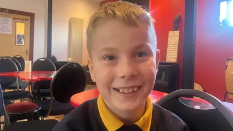 A young boy with blonde hair, he is wearing a navy jumper with yellow polo underneath