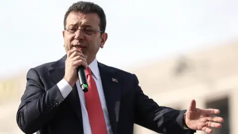 EPA EKREM IMAMOGLU, wearing a red suit and tie, carrying a microphone in his right hand and extending his left hand, and he speaks to supporters a day after the university diploma is canceled