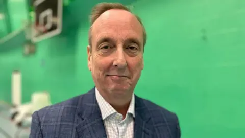 A middle aged man wearing a white checked shirt and grey checked jacket, in a sports hall