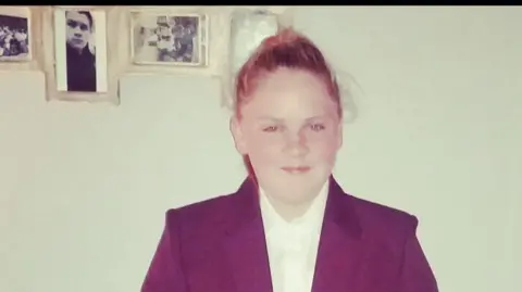Jackie Jellyman A young girl smiling at the camera in school uniform. She is wearing a maroon blazer and white shirt with an oak tree emblem on the blazer pocket. Her auburn hair is in a pony tail. 