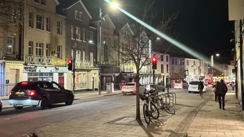 Looking down from the top of Prince of Wales Road in Norwich. Cars going down the one way street, with buildings on either sides. Traffic lights in the middle.