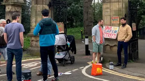 BBC Several people standing outside park next to placard that says 'save our streets'