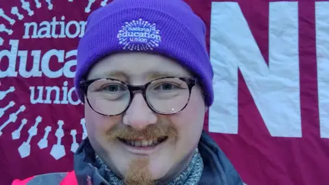 Steve Scott A man is seen smiling directly into the camera. He is wearing a pink hi-vis jacket, and has a purple hat reading National Education Union on it. He has glasses on, and distinctive facial hair.