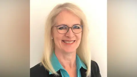 Kay Wesley A woman wearing a light blue shirt and black suit jacket is smiling at the camera. She has blonde hair and is standing against a light-coloured backdrop.