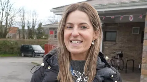 Woman with long brown hair wearing black coat. She has silver earrings. She is standing in a car park