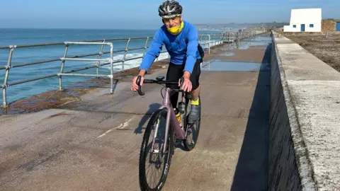 Alistair Mitchell Image shows Alistair Mitchell in a blue jumper with a yellow scarf and black cycle helmet riding his bike along a sea wall. Alistair is looking at the camera and his feet are on the pedals, hands on the handlebars. Behind and beside him the sea wall and railings are visible as is a small white building with blue doors and windows. It is a sunny day and the sea behind is relatively calm with some white water near the wall.